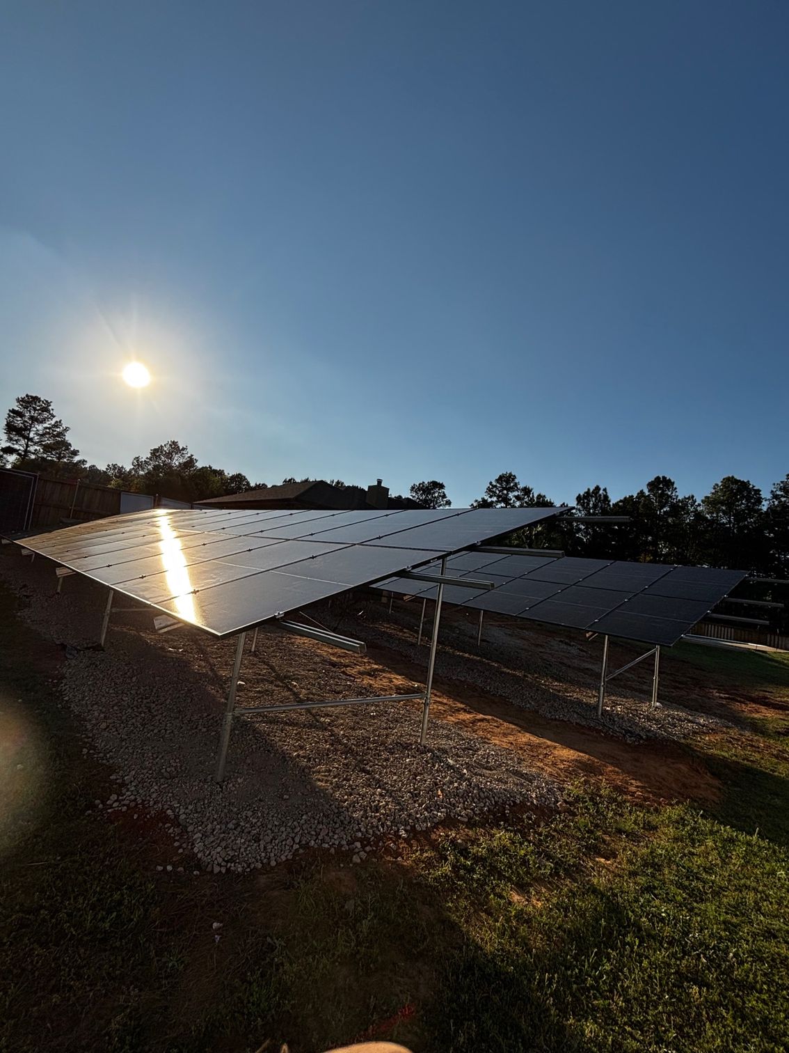 Rows of solar panels installed in a gravel-filled yard under a bright, sunny sky with trees in the distance.