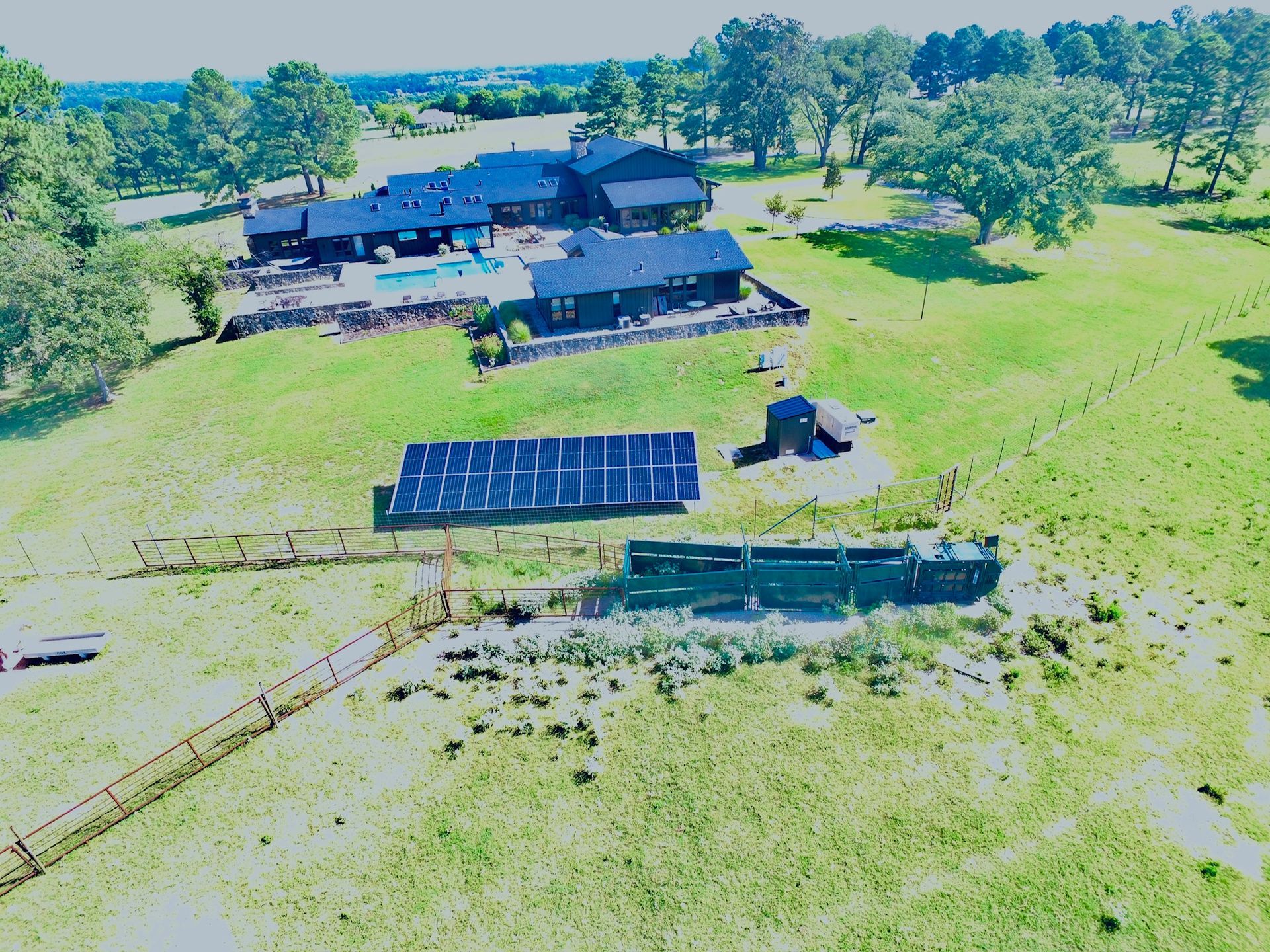 An aerial view of a dark-colored home with a pool, a large ground-mounted solar array, and a long livestock feeder.