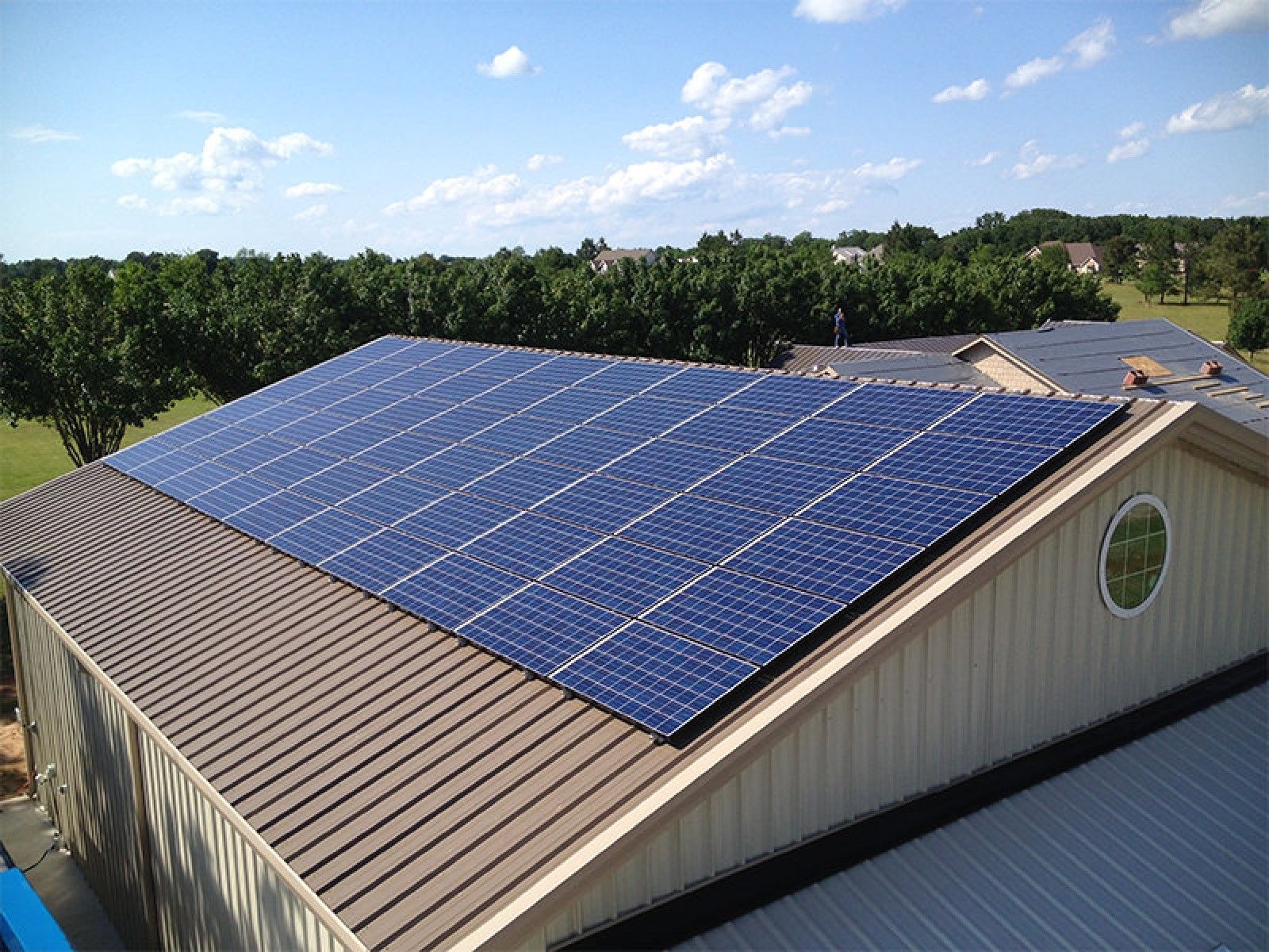A large array of dark solar panels mounted on a brown metal roof of a building, surrounded by green trees under a blue sky.
