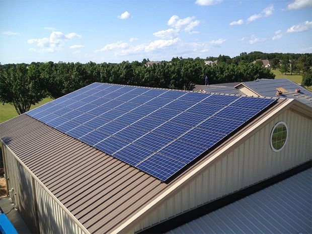 A large array of dark solar panels mounted on a brown metal roof of a building, surrounded by green trees under a blue sky.