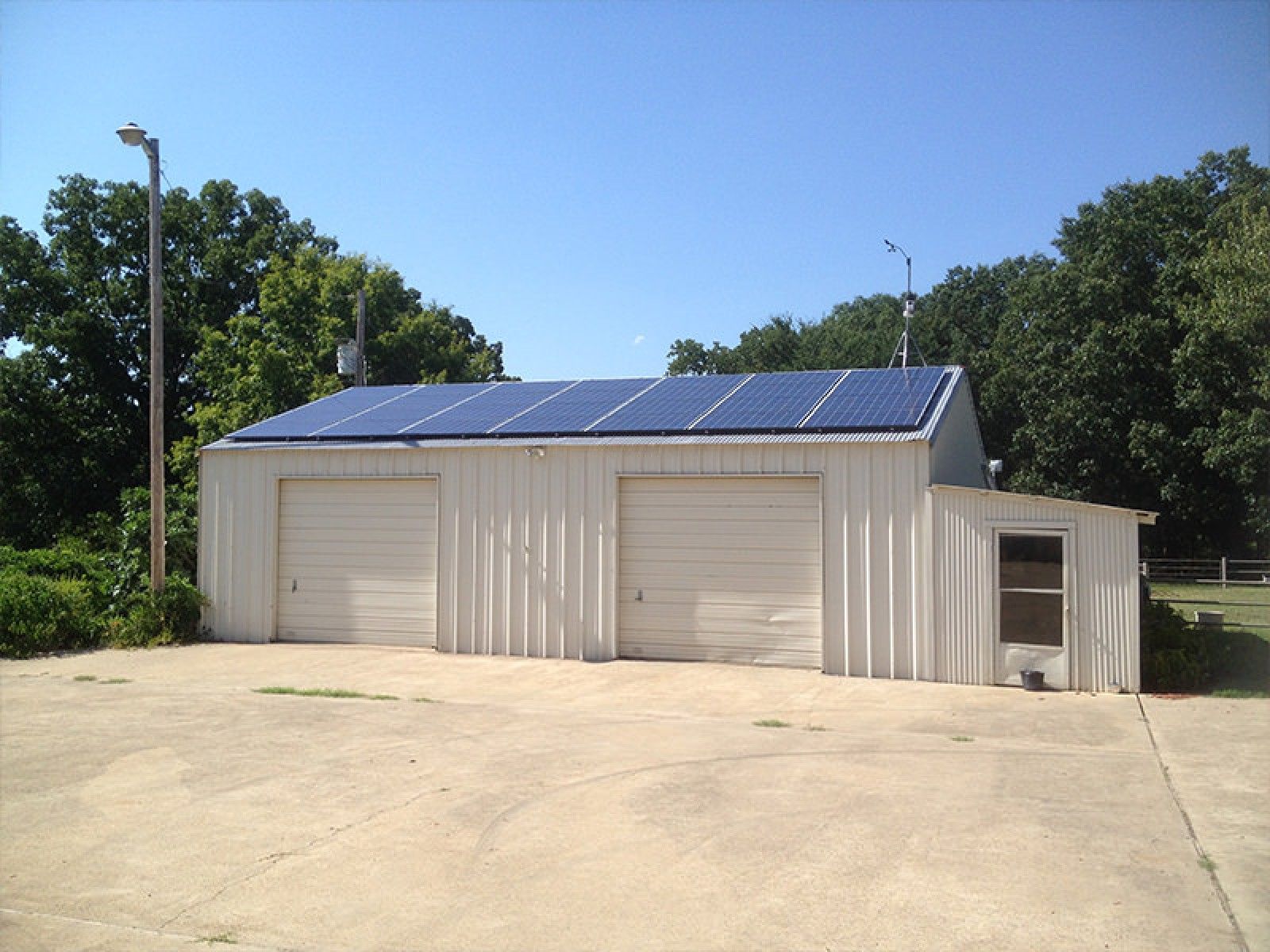 A beige metal workshop building with two garage doors and a row of solar panels on its roof, surrounded by trees.