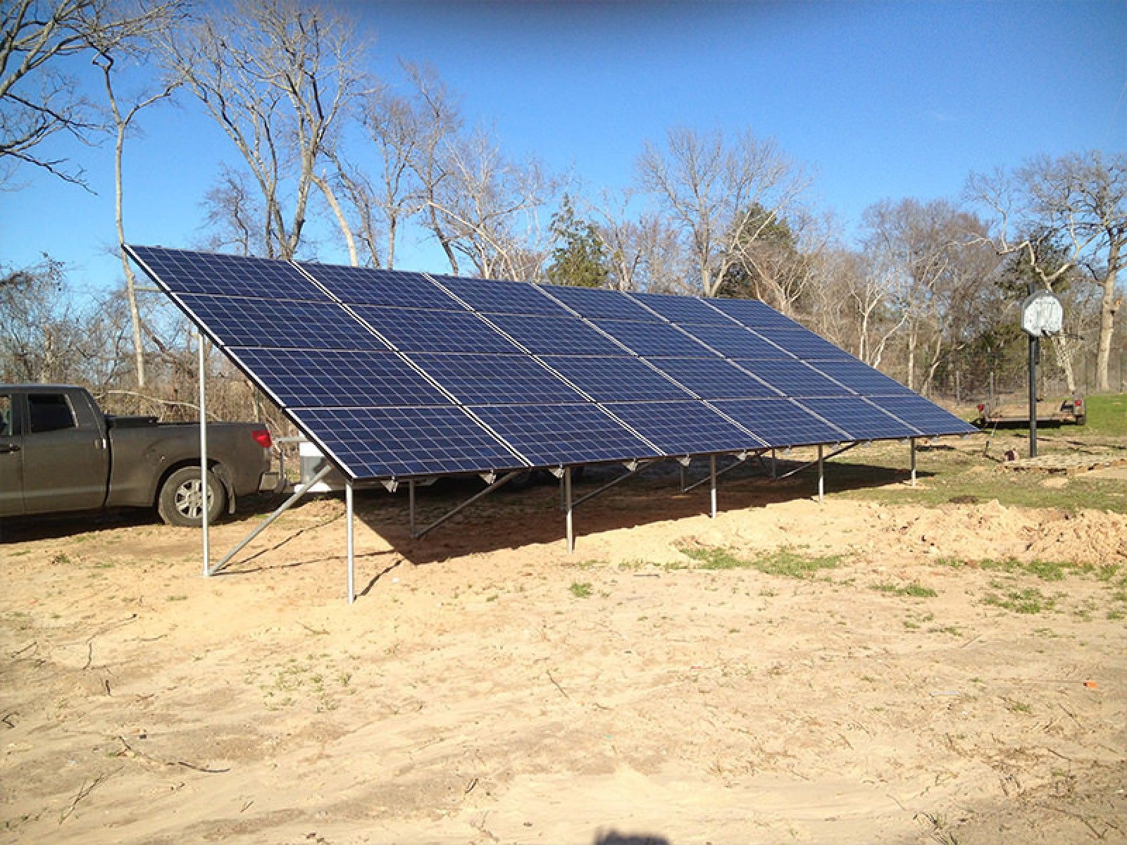 A large, ground-mounted array of solar panels installed in a sandy, outdoor clearing near a pickup truck.