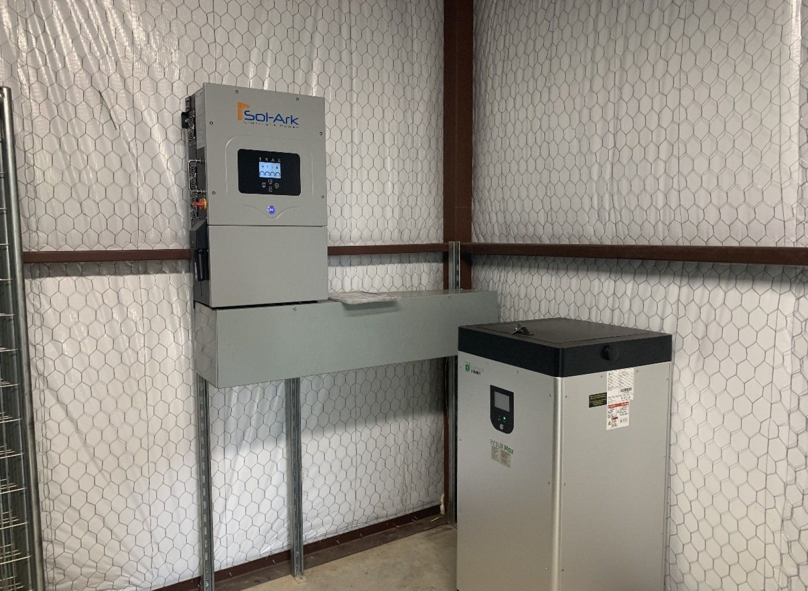 Wall-mounted solar inverter and a floor-standing battery unit in a room with white insulated walls.