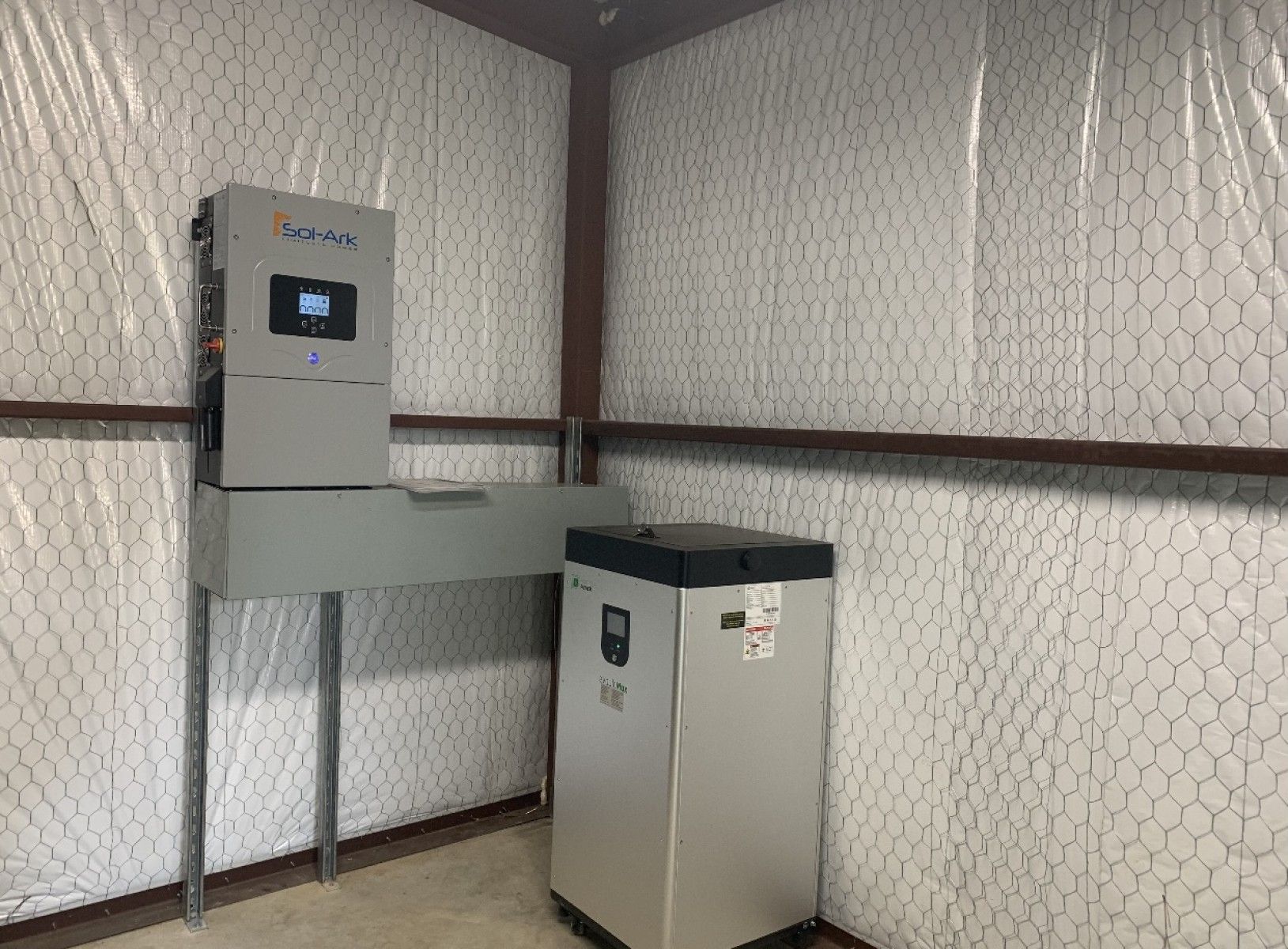 A gray solar inverter and a silver battery storage unit installed in a utility room with plastic-covered walls.