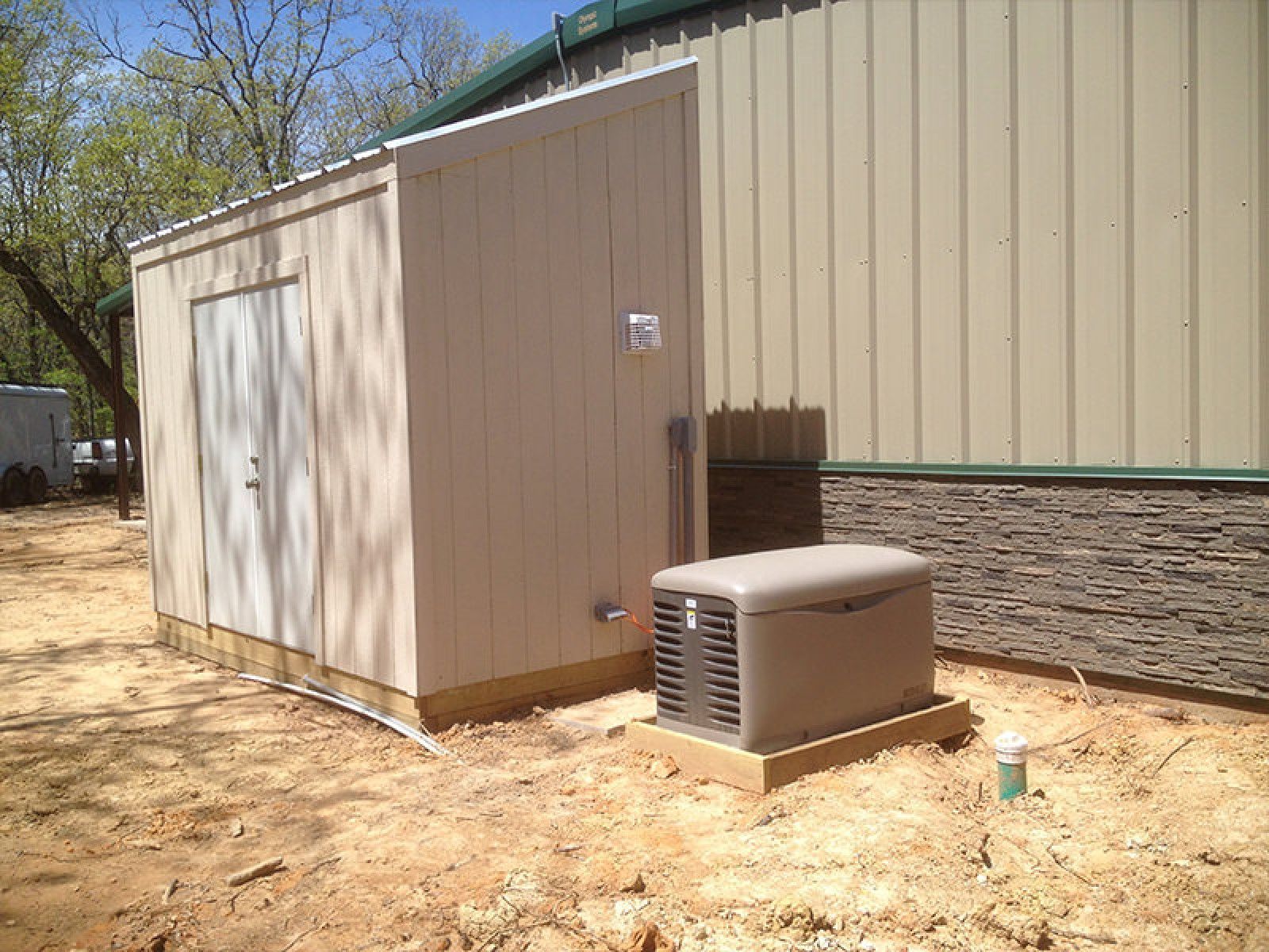 A tan shed and a standby generator sit on a sandy lot next to a tan metal building.