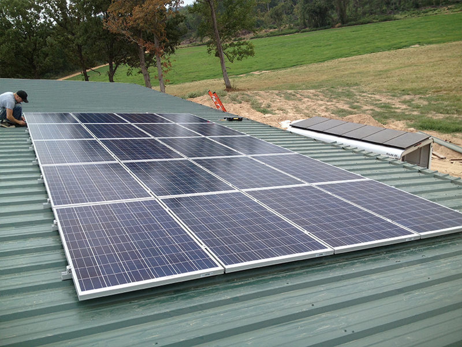 A person installing a solar panel array on a green metal roof in a rural setting.