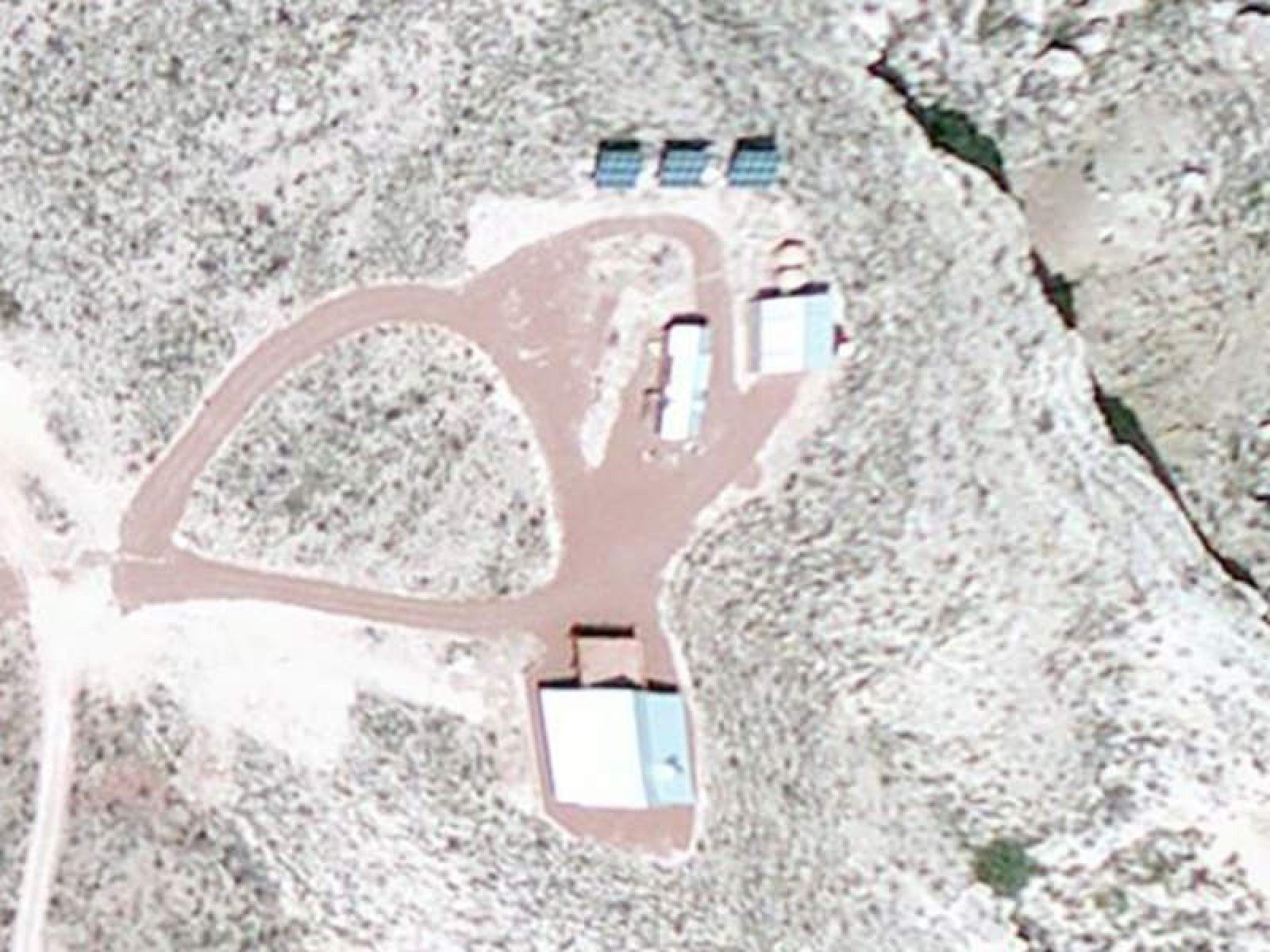Aerial view of a remote desert property with a house, several outbuildings, three solar panels, and a dirt driveway.