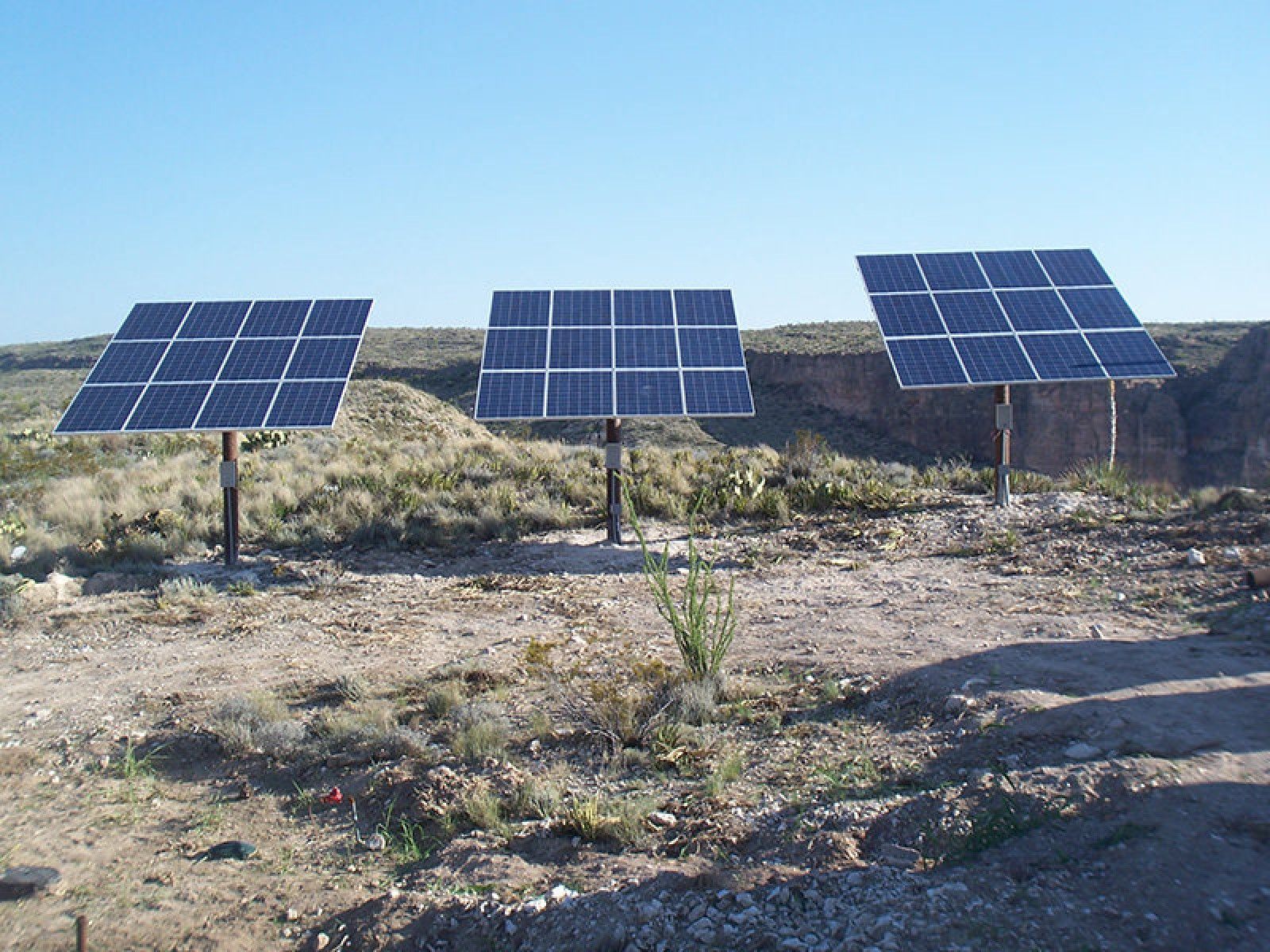 Three solar panel arrays stand on poles in a dry, desert landscape under a clear blue sky.