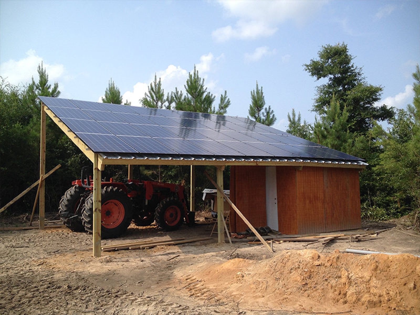 A large solar panel array mounted on the sloped roof of a wooden equipment shed, with a red tractor parked underneath.