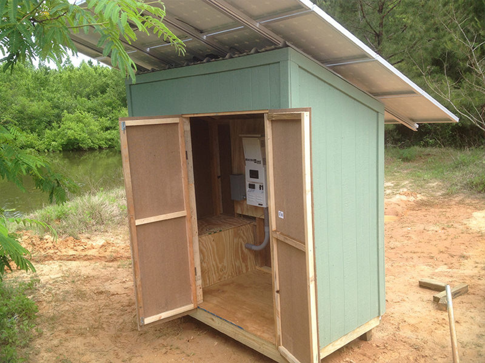A small, light green wooden shed with open double doors stands outdoors, featuring a solar panel mounted on its roof.