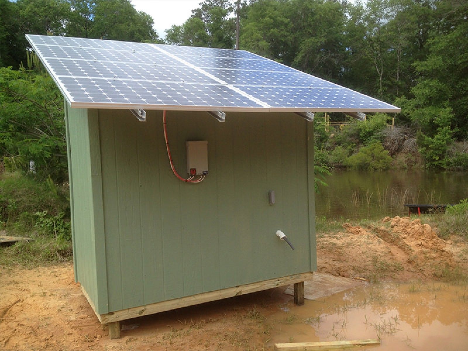 Two solar panels mounted on the slanted roof of a small green shed near a pond.