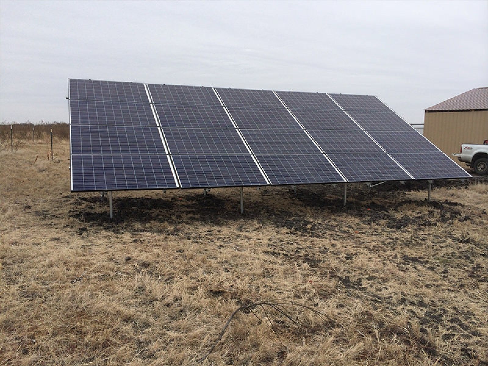 A ground-mounted solar panel array stands in a field under an overcast sky, with a building visible to the right.