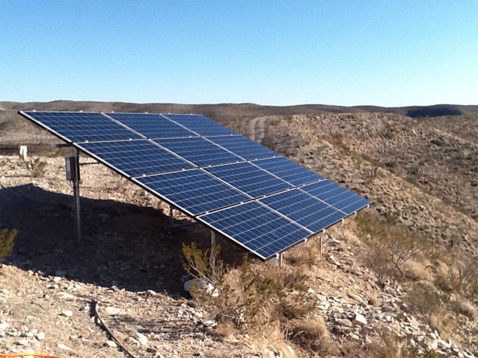 A ground-mounted solar panel array sits on a rocky, desert hillside under a clear blue sky.