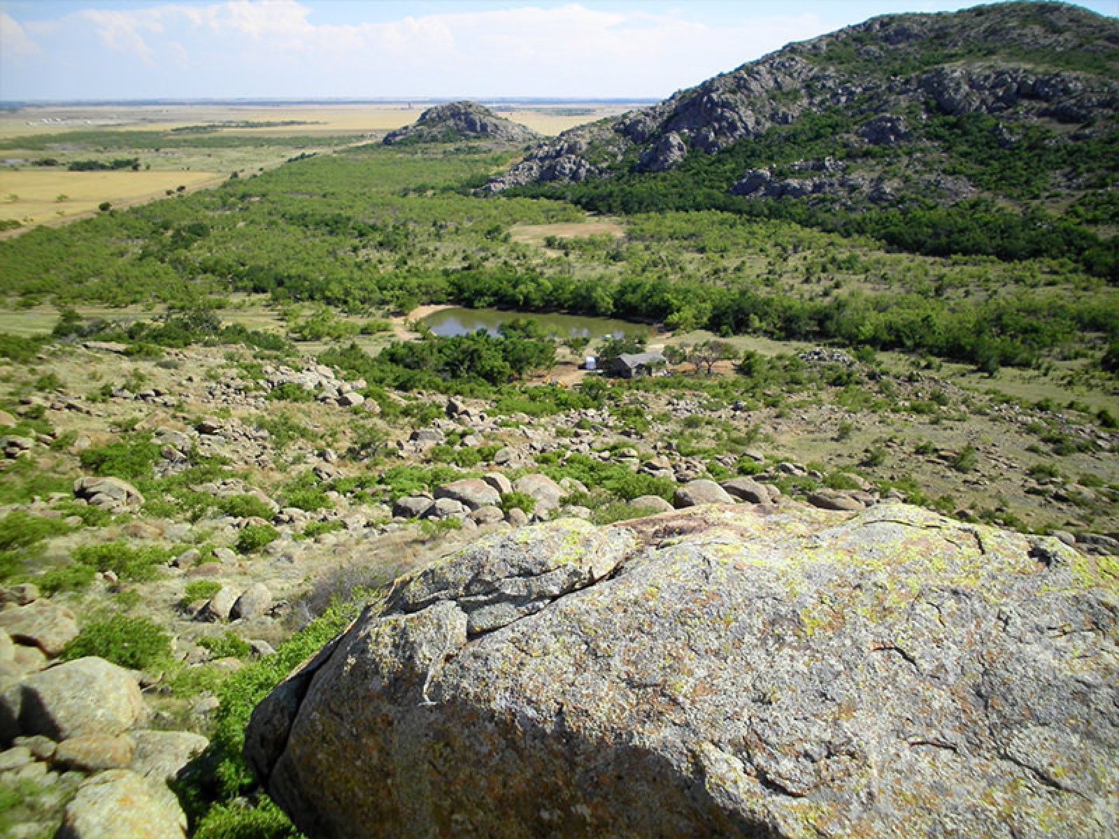 A large boulder sits in the foreground, overlooking a valley with trees, a small pond, and distant rolling hills.