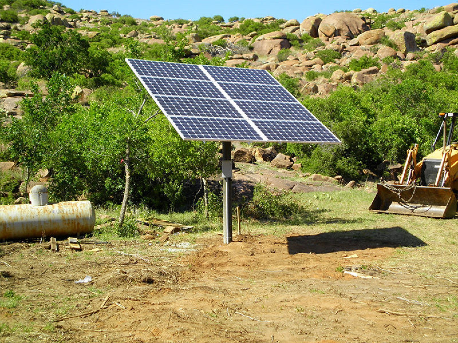 A solar panel array mounted on a single metal pole in a rocky, rural landscape with a piece of heavy machinery nearby.