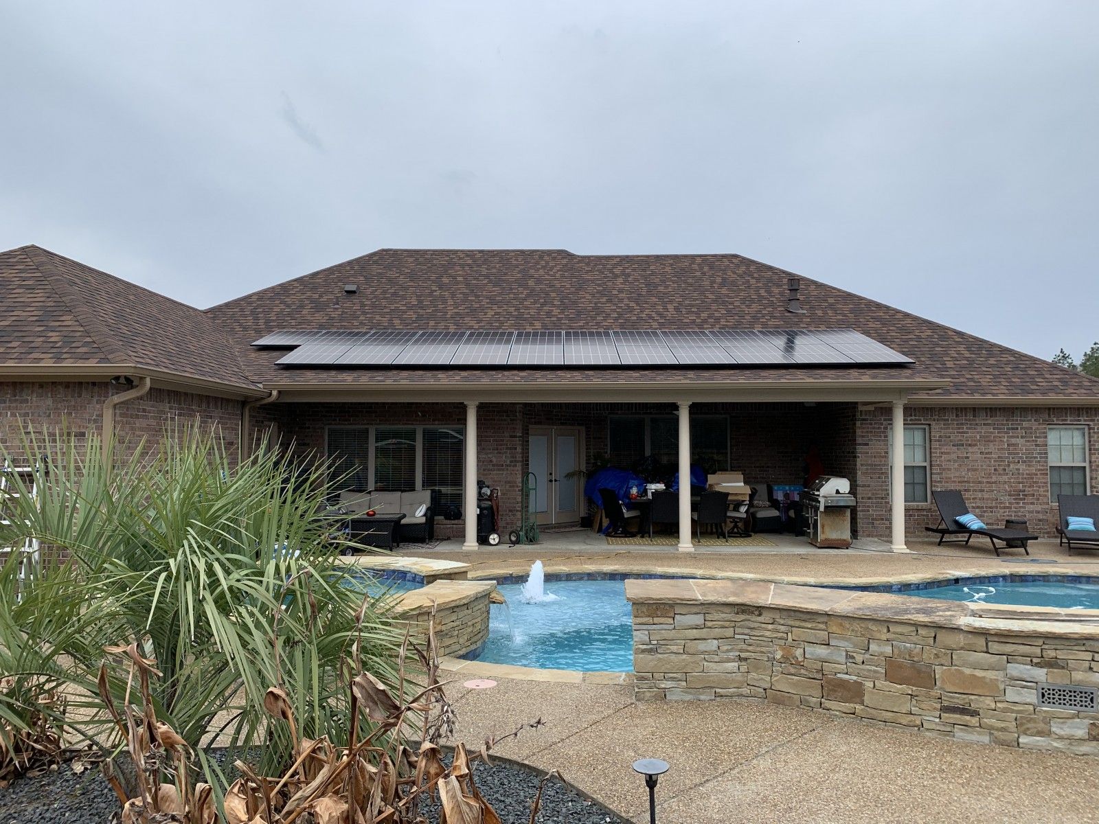 A backyard with a swimming pool and fountain in front of a brick house featuring solar panels on the roof.