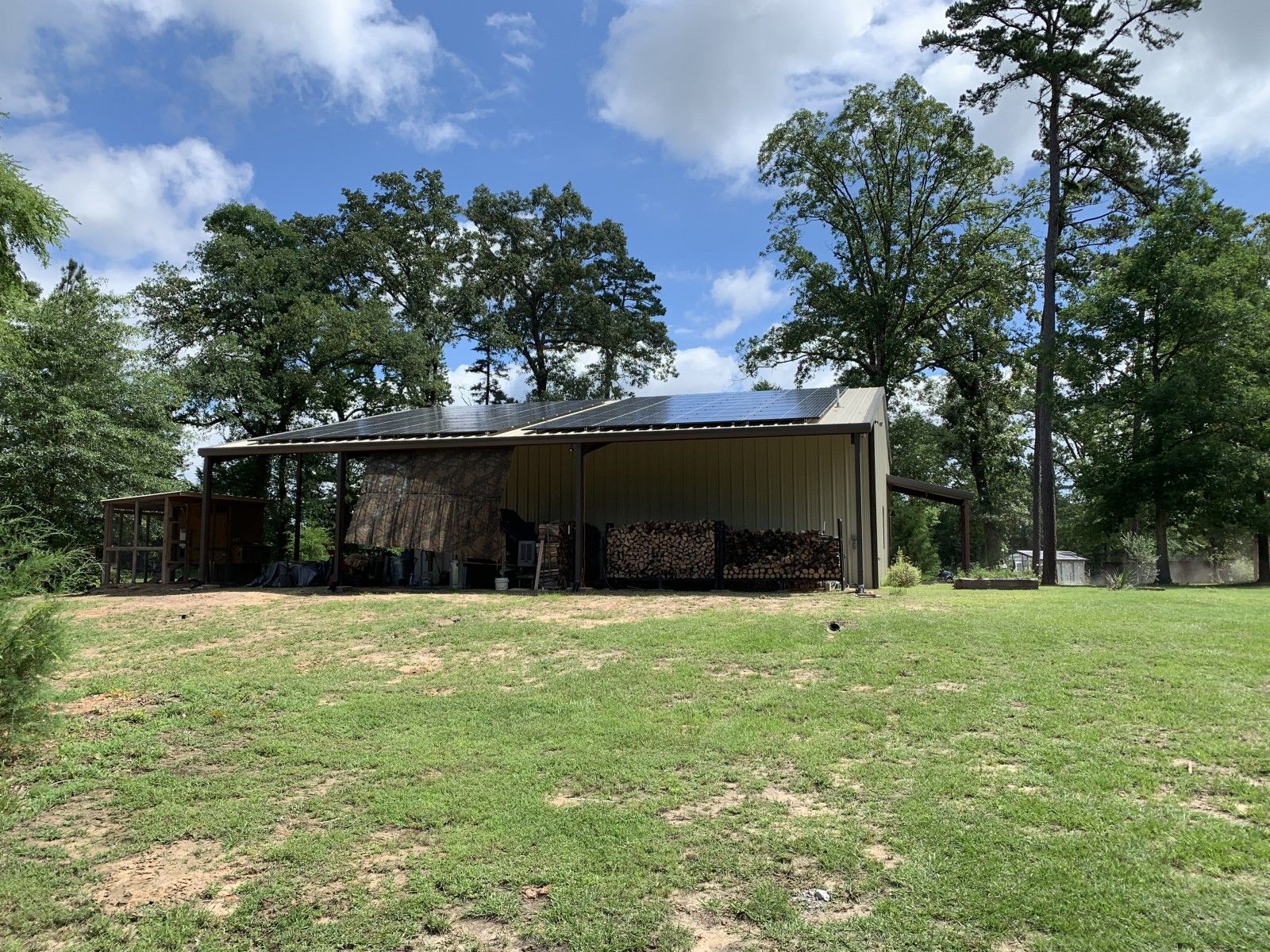 A tan metal shed with a slanted roof sits on a grassy lawn, with stacked firewood under a covered, open-air section.