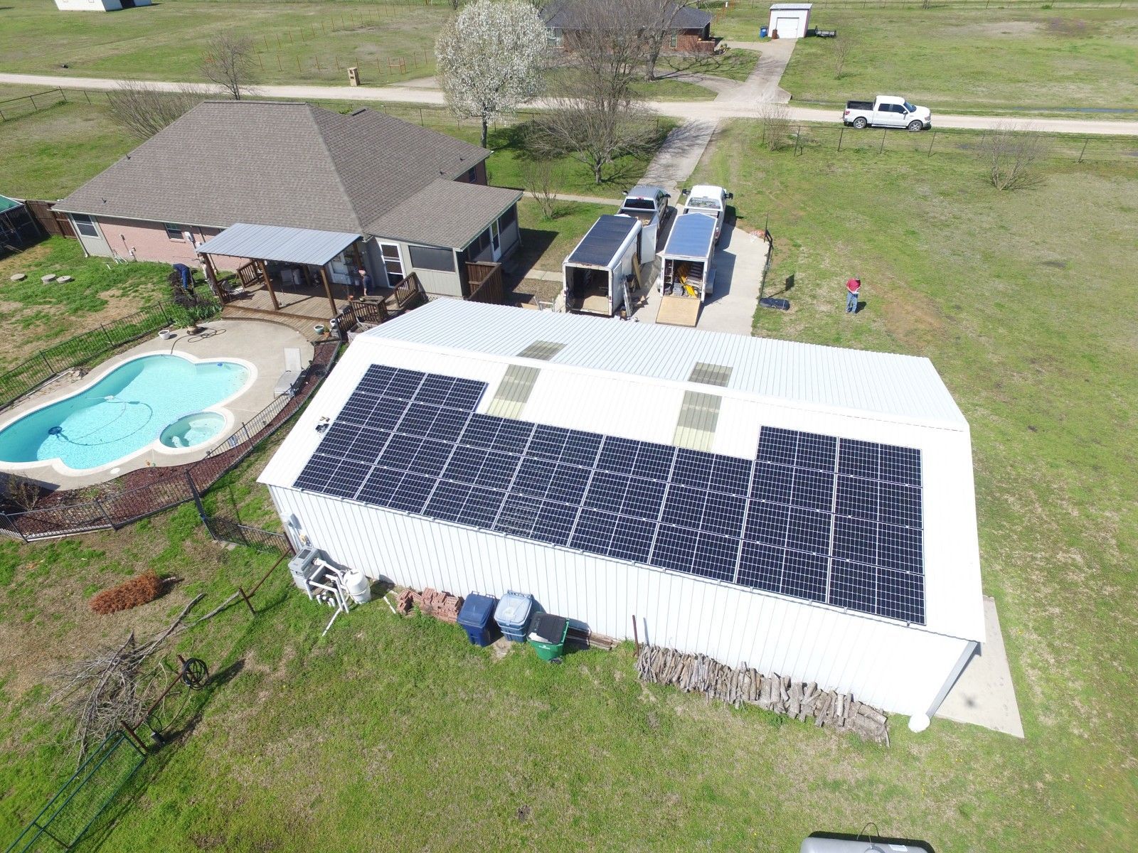 An aerial view of a white outbuilding with a large solar panel array on its roof, near a house with a swimming pool.