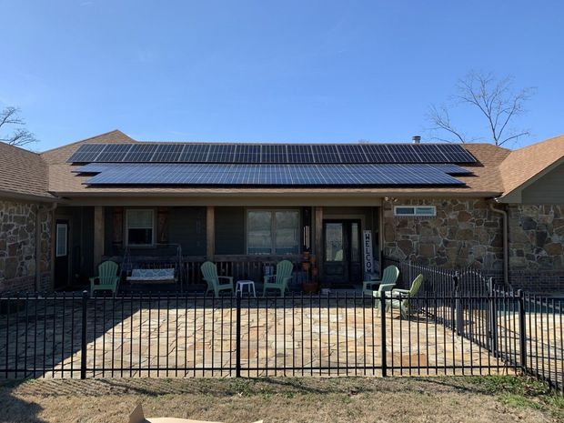 A house with a large solar panel array on the roof, a stone exterior, and a fenced-in stone patio with lawn chairs.