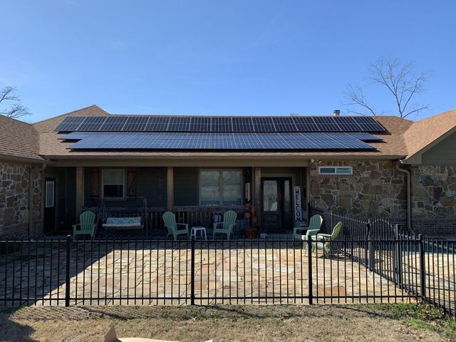 A house with a large solar panel array on the roof, a stone exterior, and a fenced-in stone patio with lawn chairs.