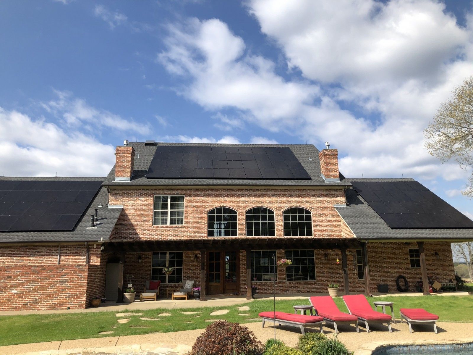A brick home with solar panels installed on the roof, featuring a patio with lounge chairs and a grassy yard.