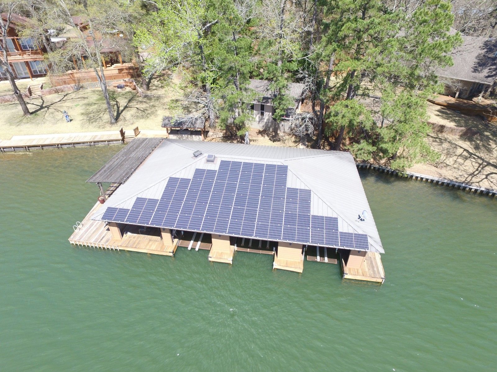 An aerial view of a boat dock on a lake with a large array of solar panels mounted on its flat roof.