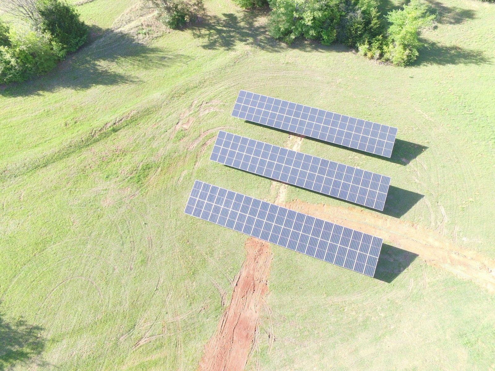 An aerial view of three long rows of solar panels situated in a grassy field with a dirt path running between them.