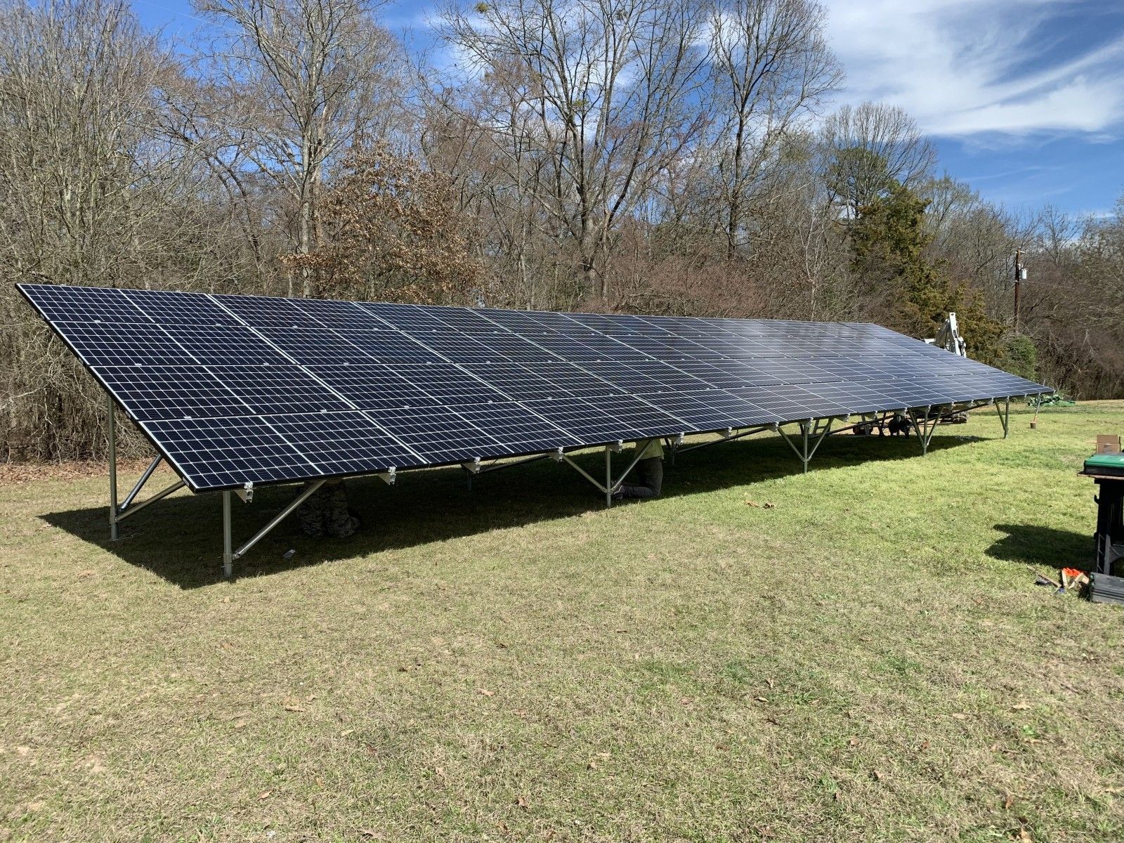 A large, ground-mounted solar panel array installed in a grassy field with trees in the background under a blue sky.