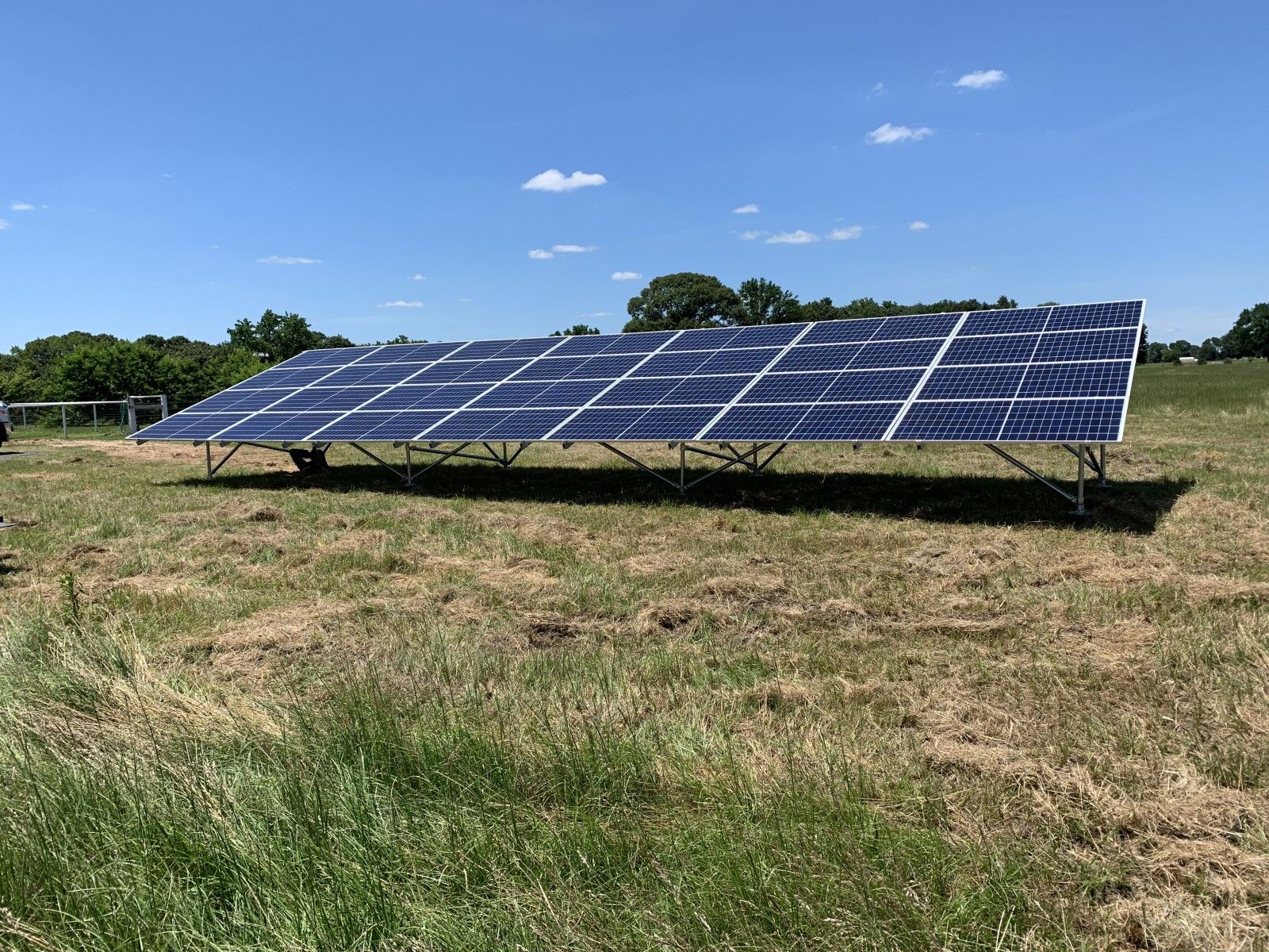 A ground-mounted solar panel array stands in a grassy field under a bright blue sky.