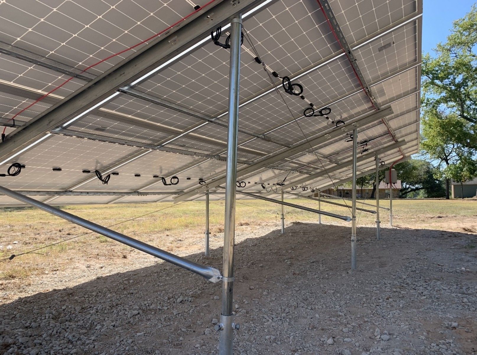 A low-angle view looking beneath a ground-mounted solar panel array installed on a gravel field under a clear blue sky.