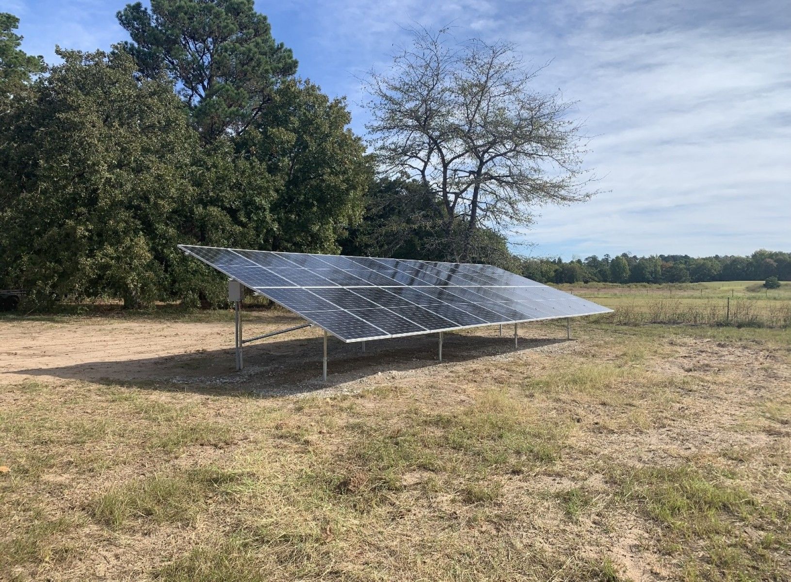 A solar panel array mounted on a metal frame sits in a grassy field in front of a tree line under a partly cloudy sky.