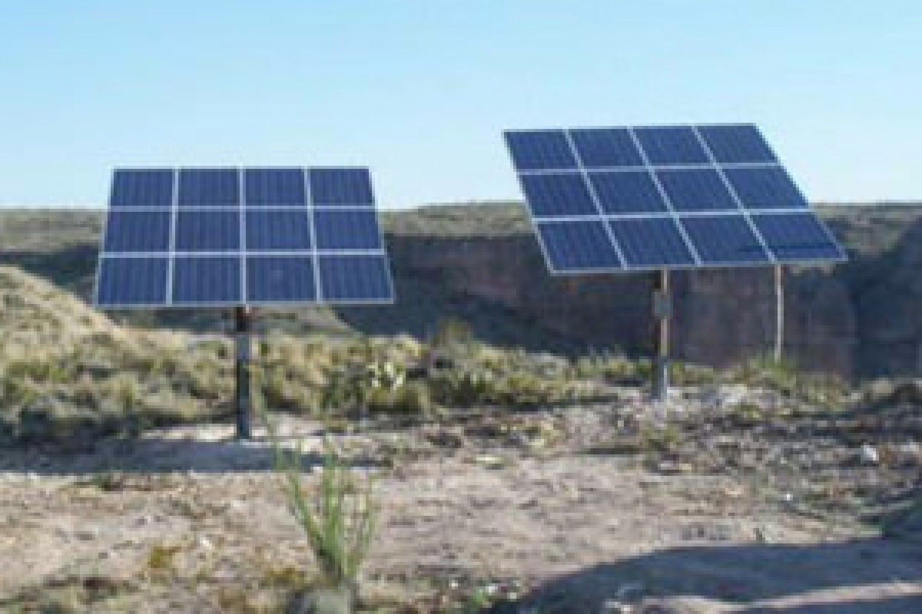 Two solar panel arrays stand on a dirt hillside against a blue sky, overlooking a rocky canyon.