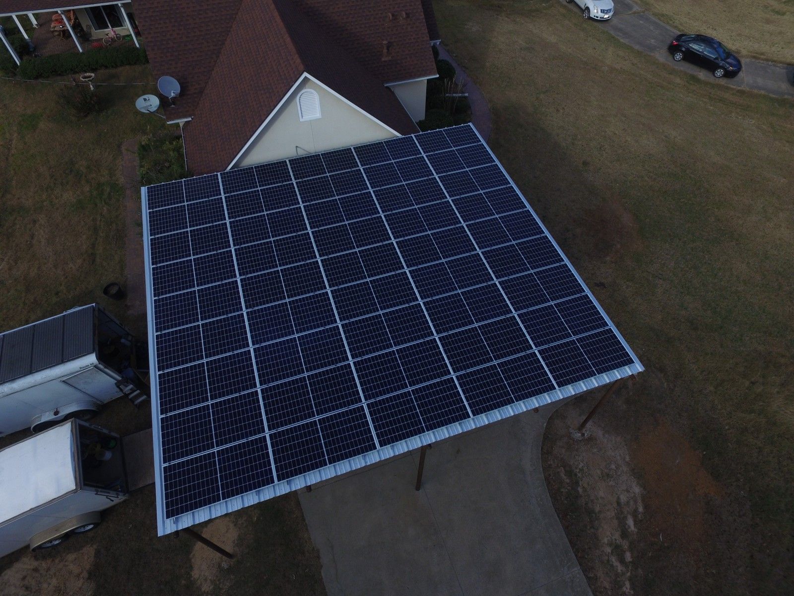 An aerial view of a large, square solar panel array mounted on a carport structure next to a house.