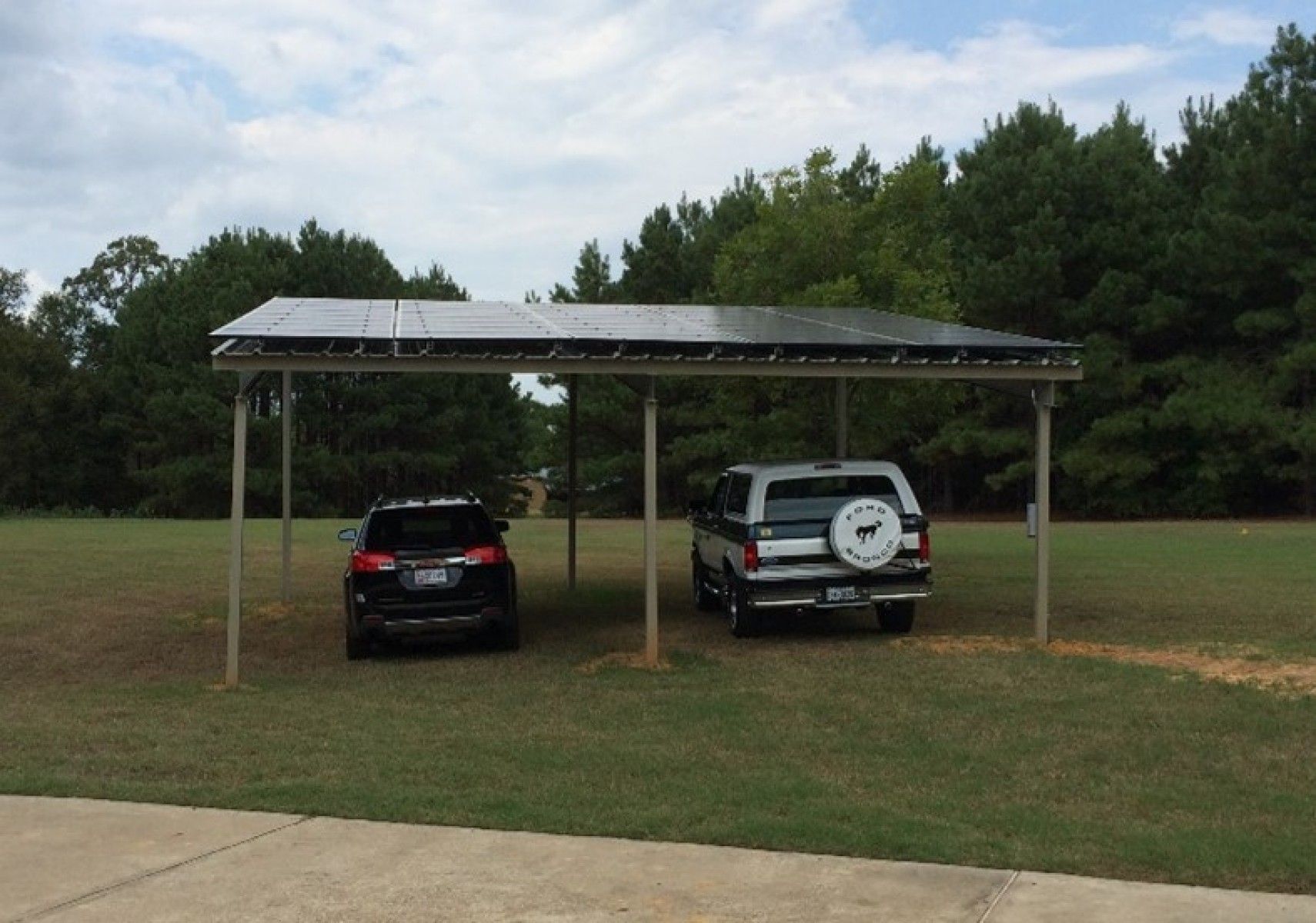 Two cars parked under a rectangular solar panel carport in a grassy field with trees in the background.