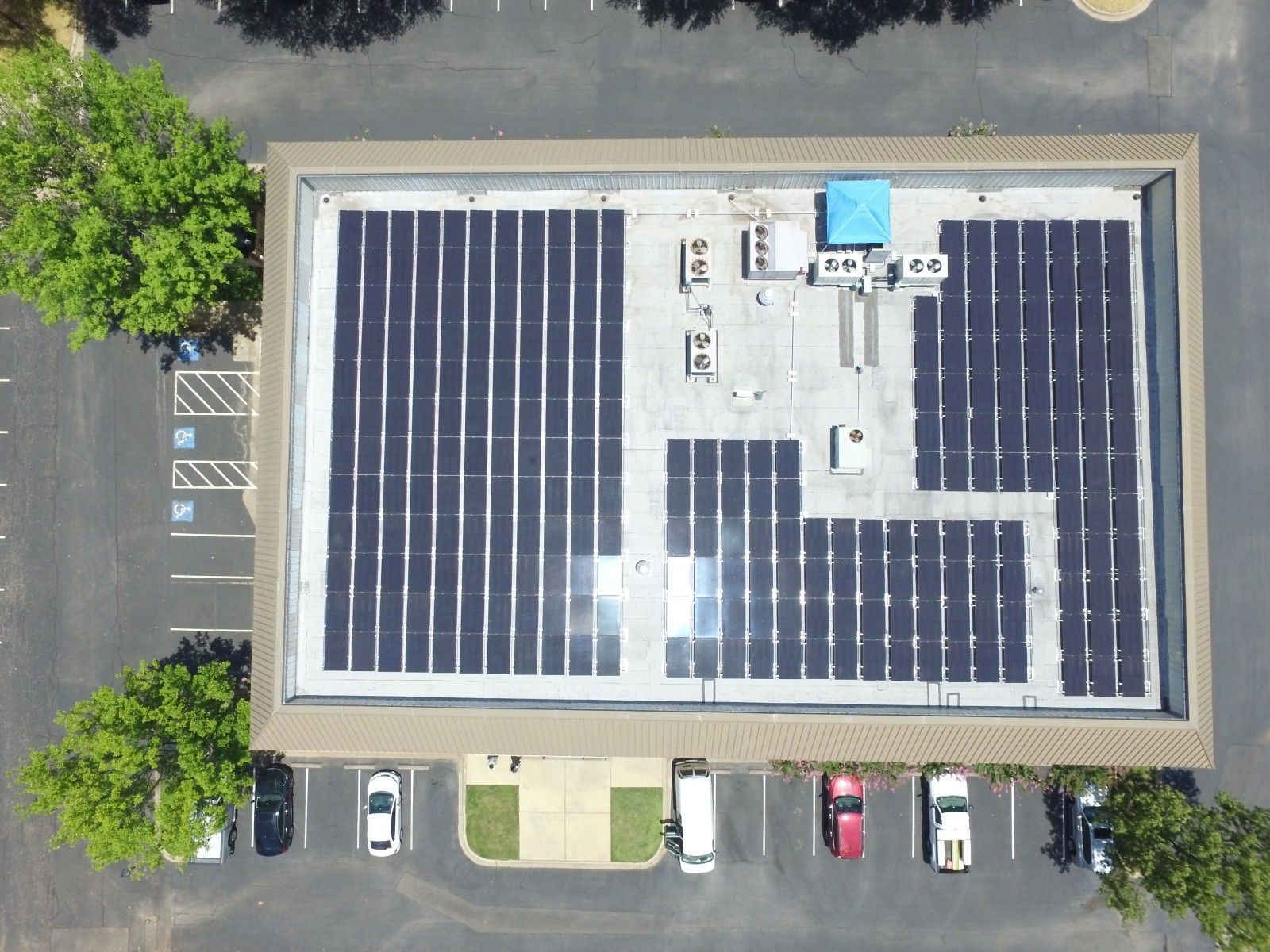 An aerial view of a flat commercial roof covered in rectangular black solar panels, with a parking lot below.