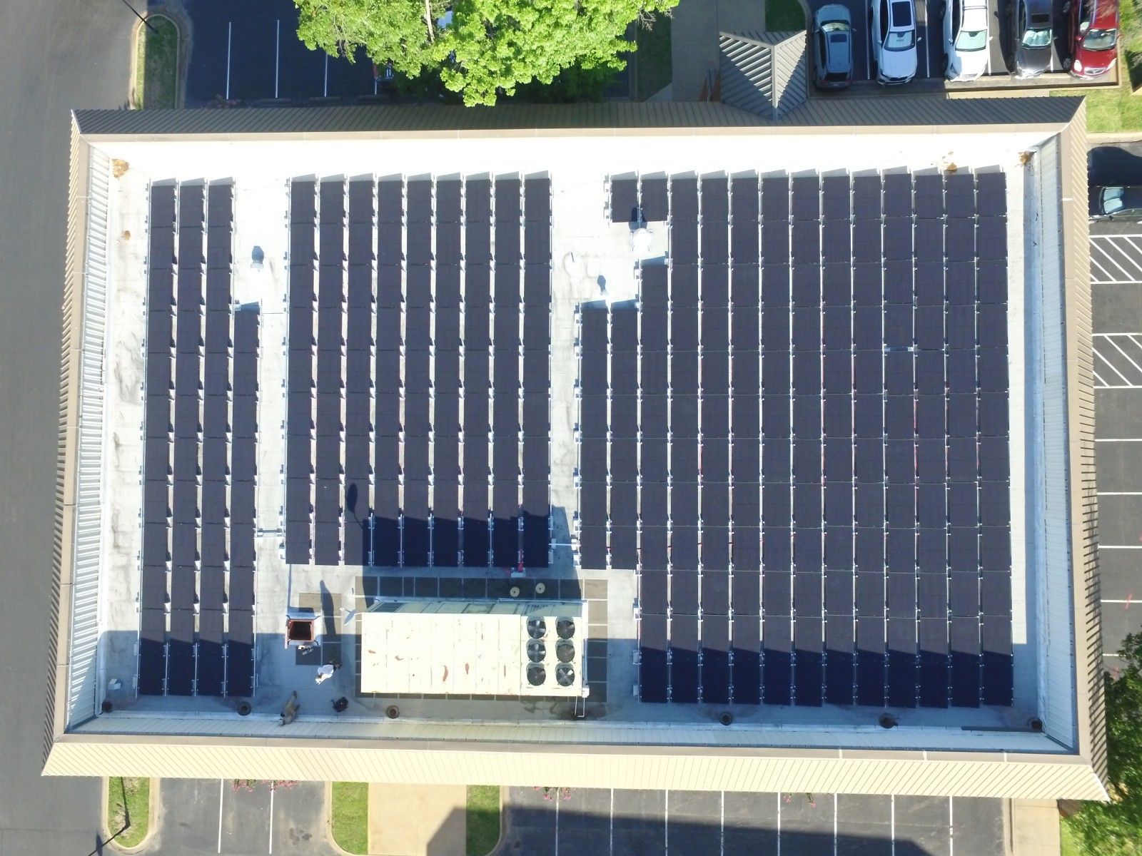 Aerial view of a flat building roof covered with rows of solar panels, featuring a central mechanical unit.