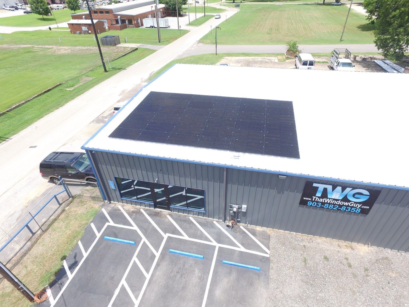 Aerial view of a gray commercial building with solar panels on the roof and a parking lot with marked spaces.