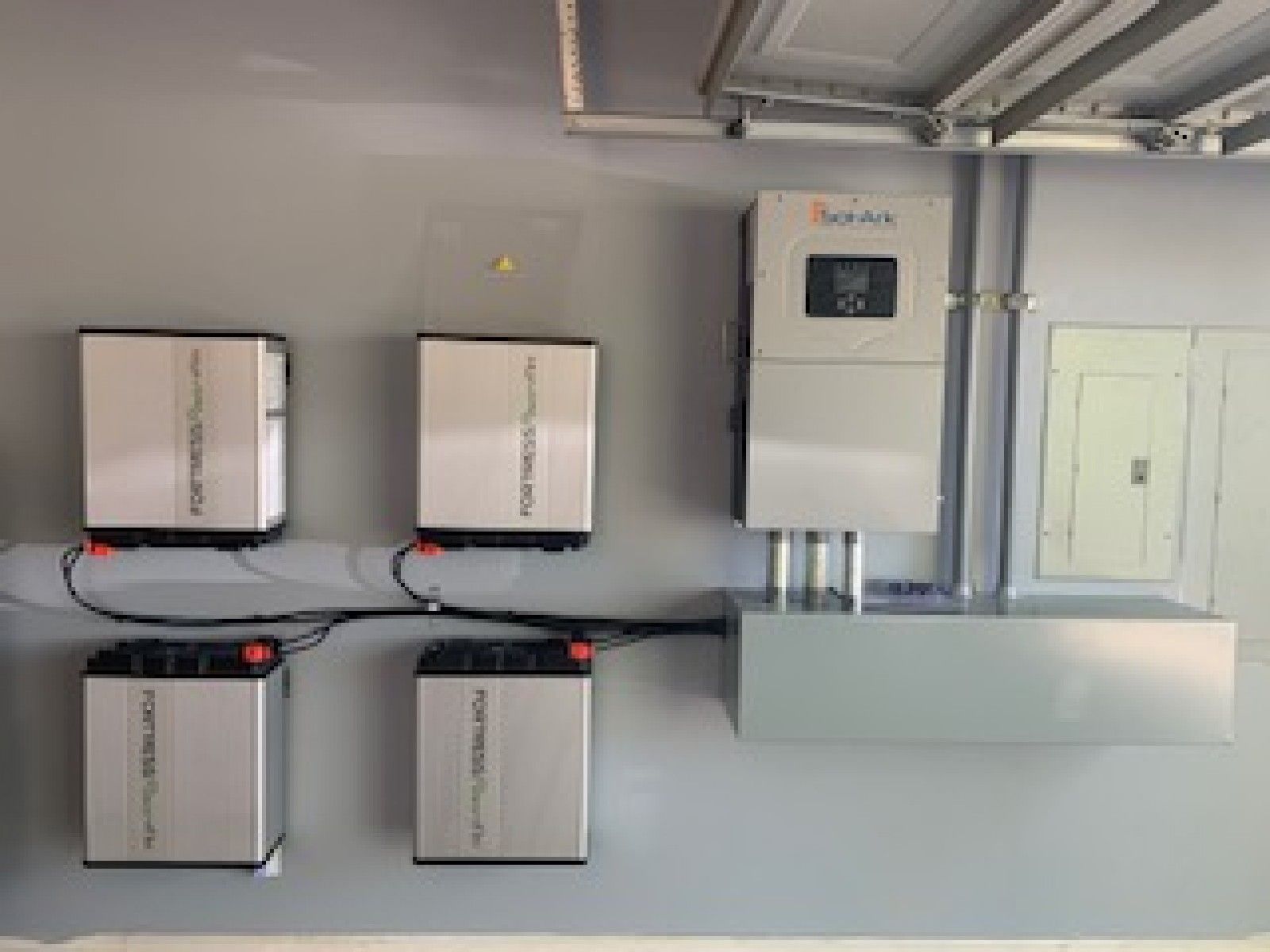 Four silver Fortress Power batteries connected to a Sol-Ark inverter and electrical panel mounted on a gray garage wall.