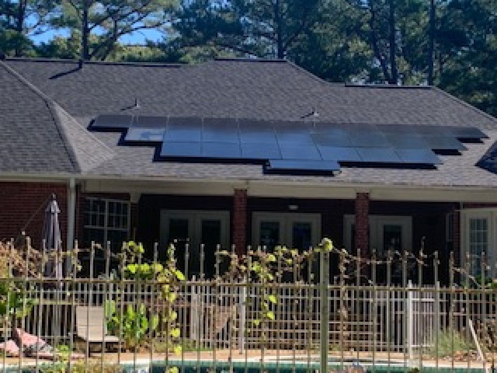 A residential house with a large array of black solar panels installed on the dark shingled roof above a backyard patio.
