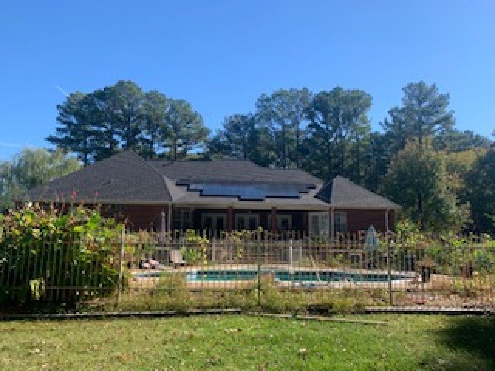 A red brick house with a dark roof and solar panels, situated behind a swimming pool, a metal fence, and a green yard.