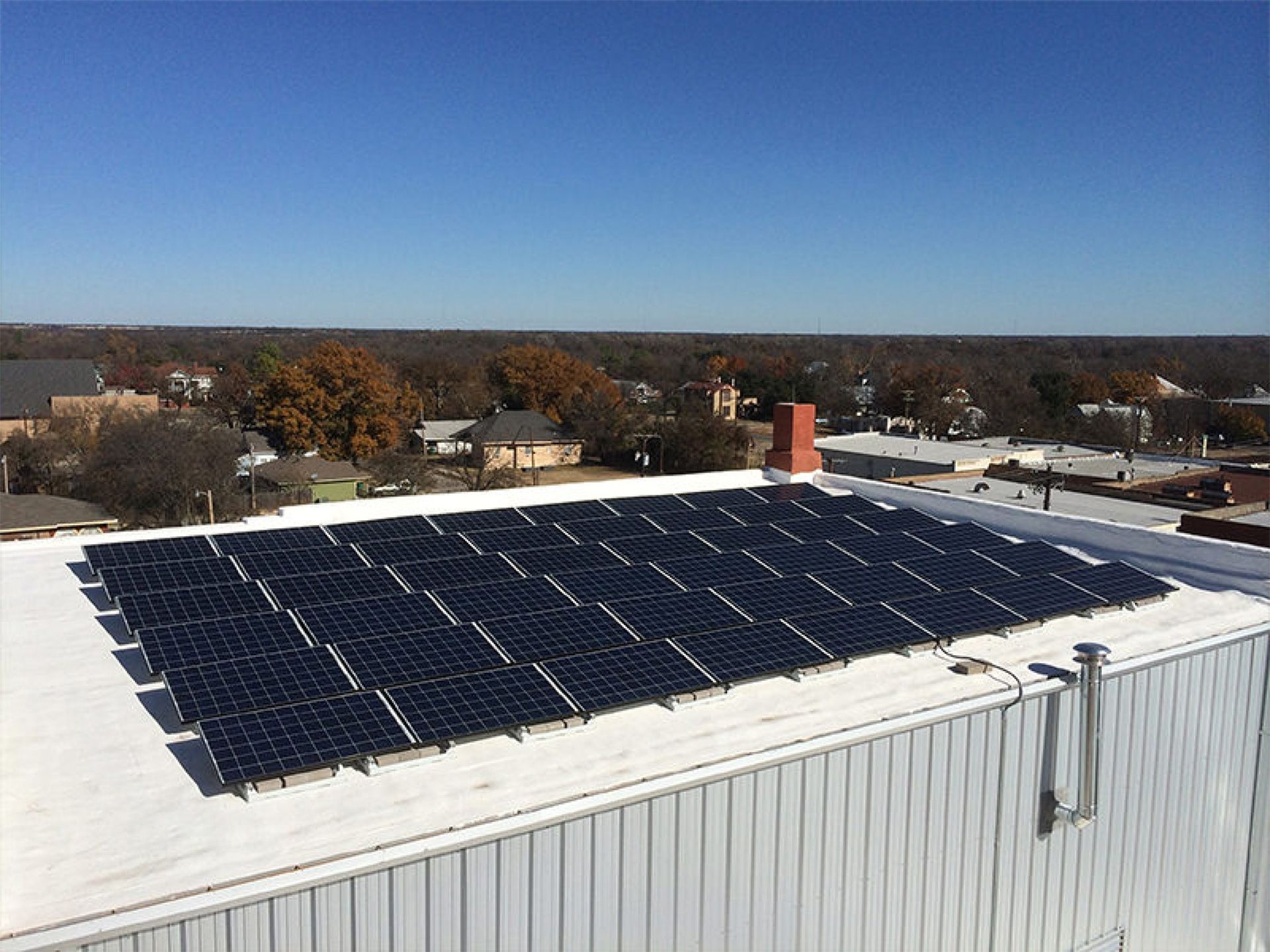 Solar panels on a white rooftop under a clear blue sky, with houses and trees in the background.