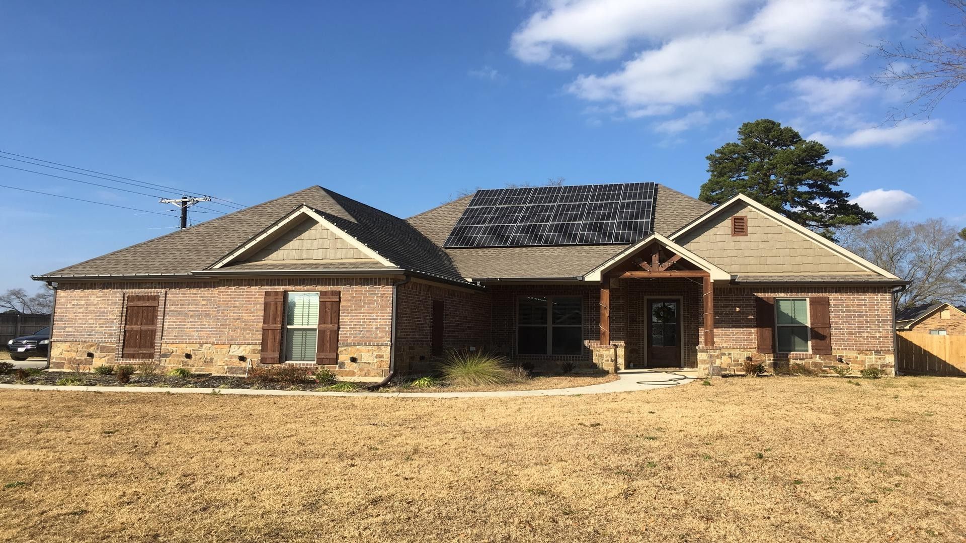 A single-story brick house with a brown shingle roof, solar panels, and a front porch under a clear blue sky.