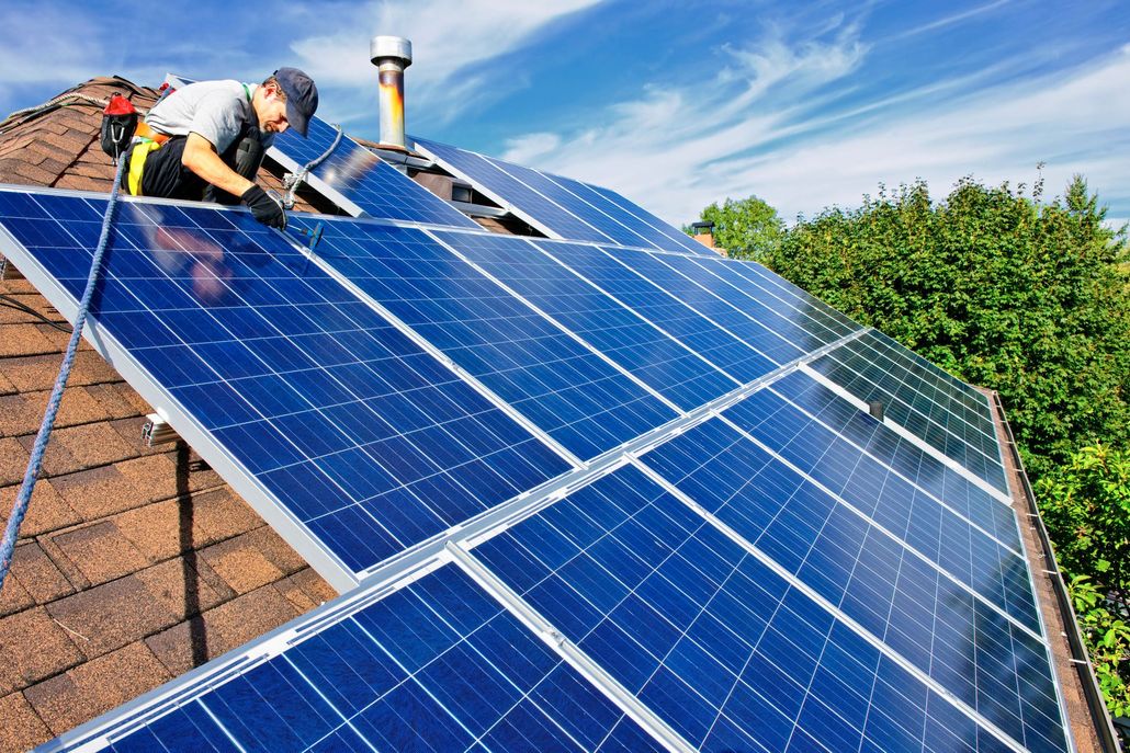 Worker installing blue solar panels on a sloped roof under a clear sky
