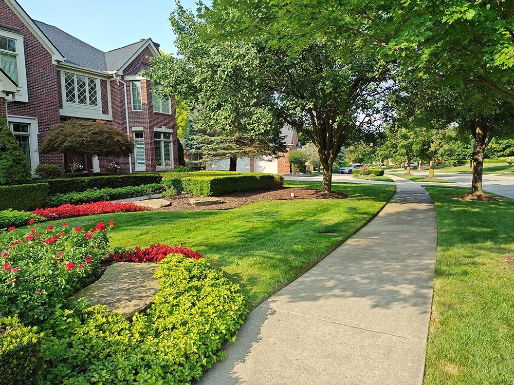 Curved sidewalk beside red-brick townhouses, green lawns, and colorful flower beds on a sunny day