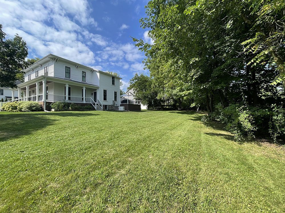 White two-story house with a porch beside a large green lawn under a blue sky.