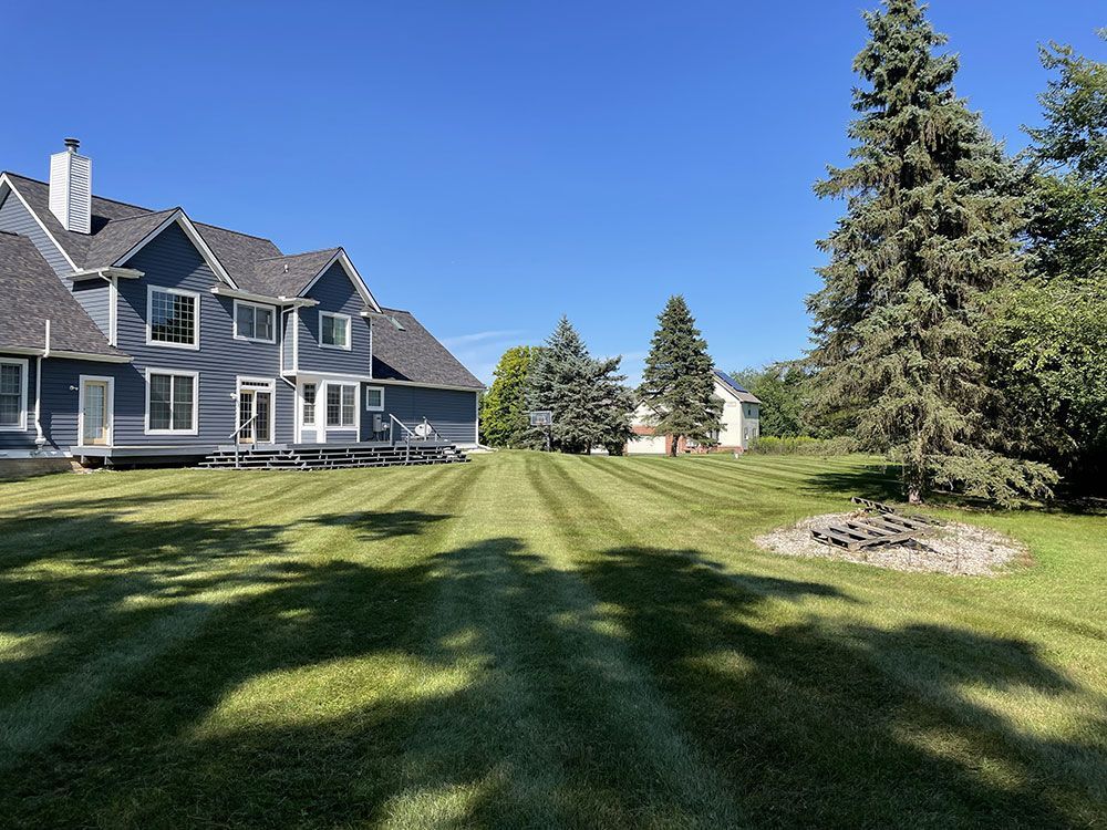 Large blue-gray house with a manicured lawn and tall trees under a clear blue sky