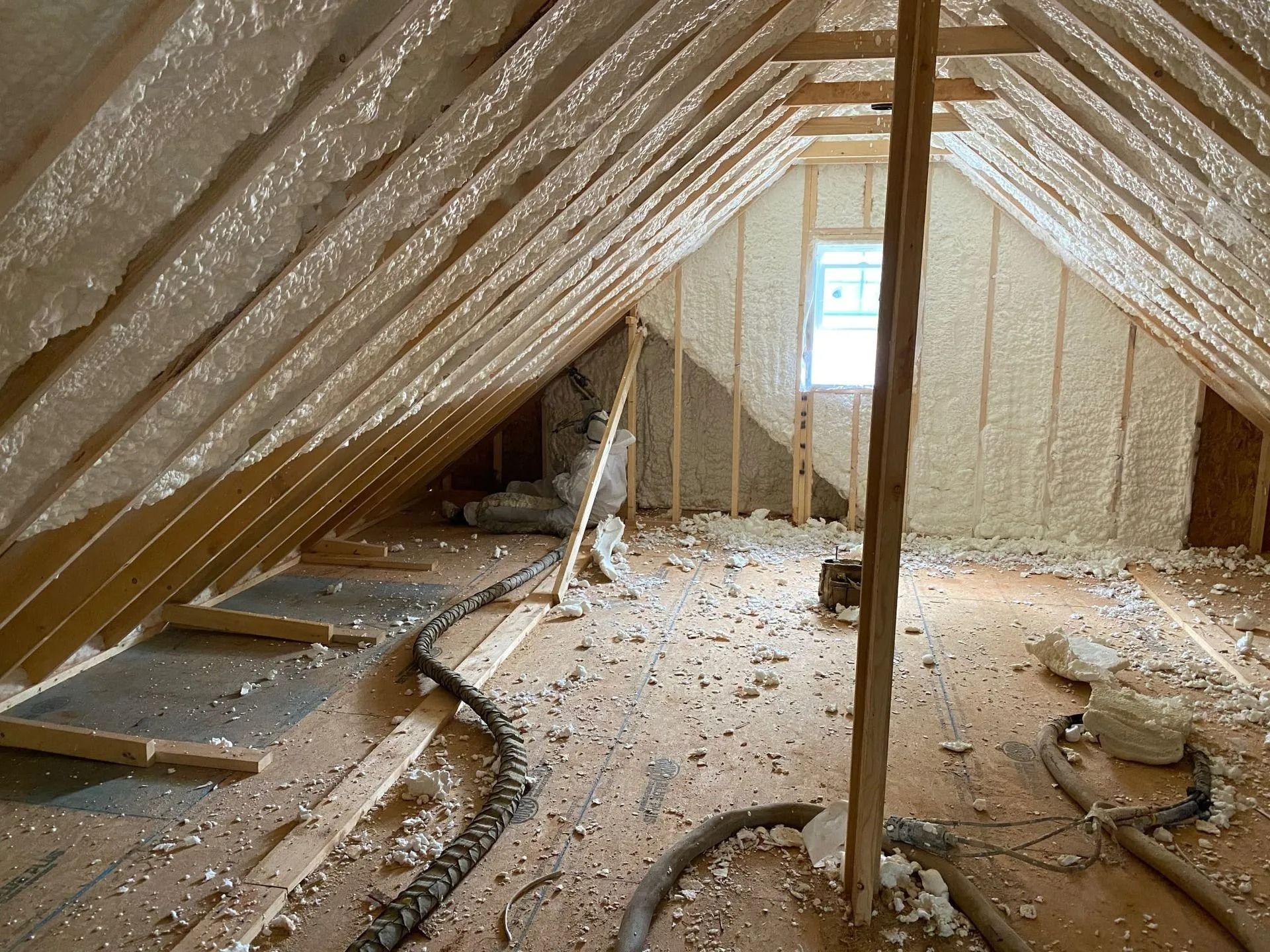 Attic interior with spray foam insulation on roof and walls, debris on floor, and window in the distance.