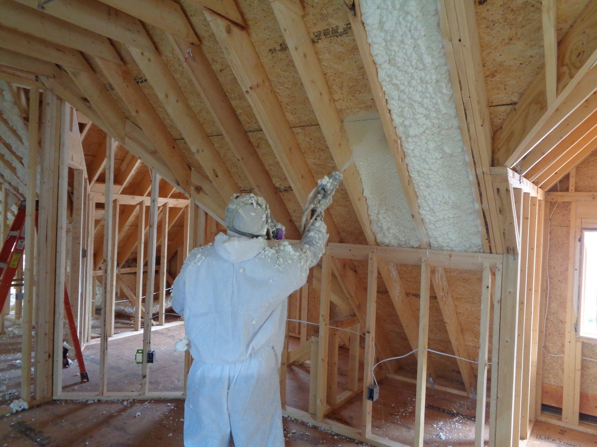 Person in protective suit spraying insulation foam inside a wooden house frame.