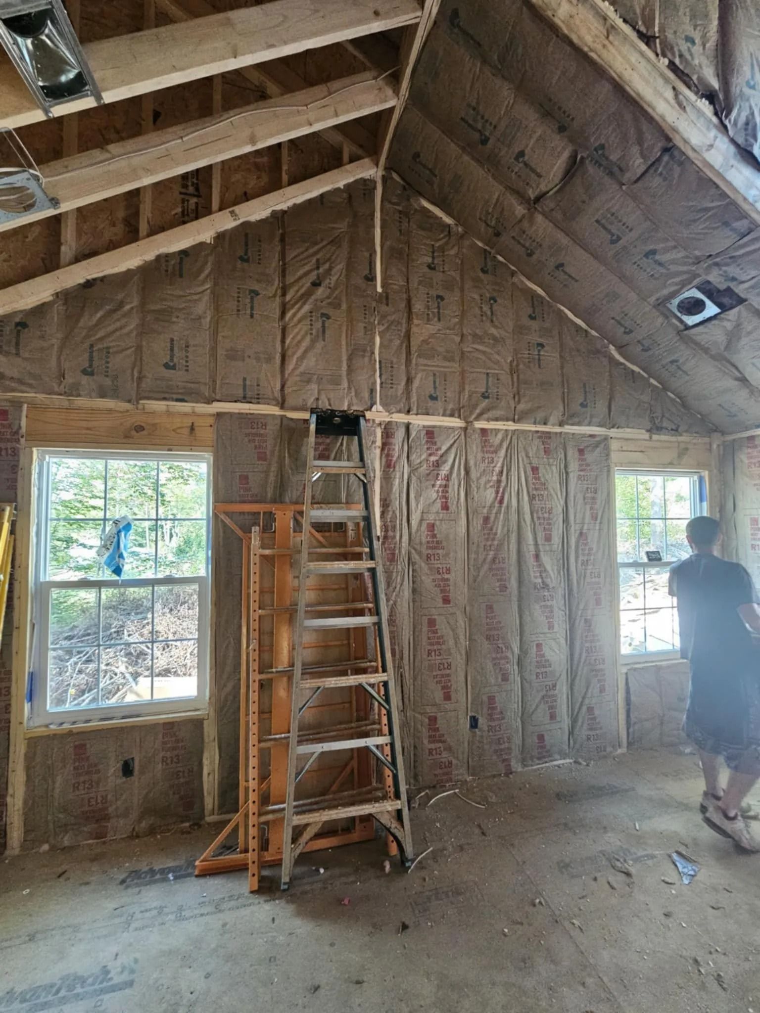 Interior of a building under construction. Walls are insulated; windows and ladder are visible. A person stands near a window.