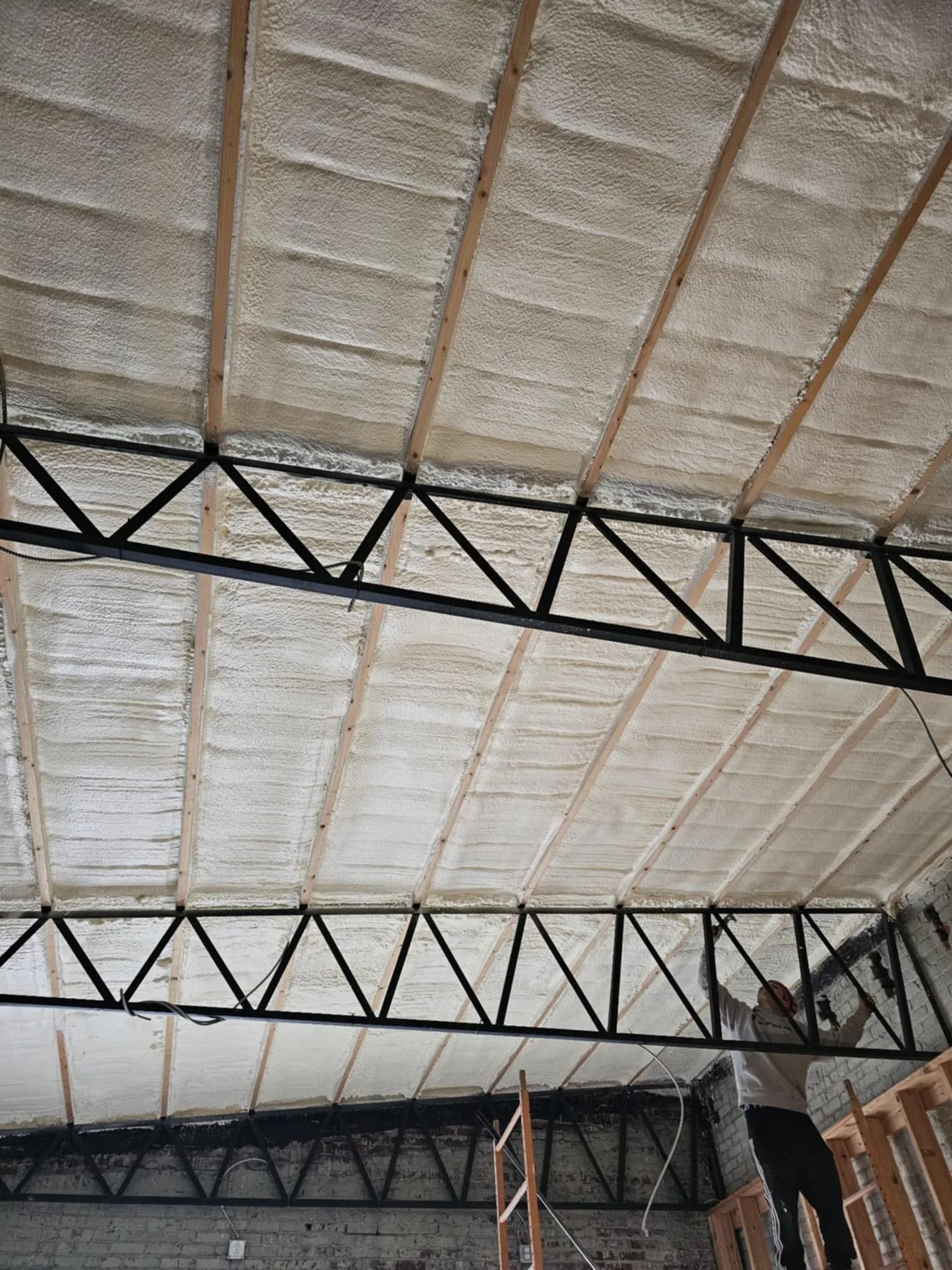 Person working on a ceiling insulated with spray foam, beneath wooden beams and metal trusses.